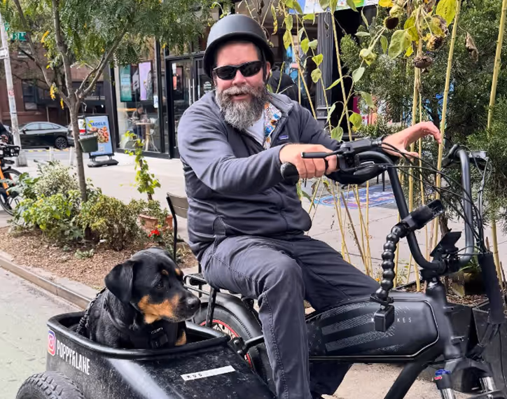 Bearded man wearing a helmet and sunglasses rides a black electric tricycle with a Rottweiler dog seated in the attached sidecar labeled PoppyFlare.